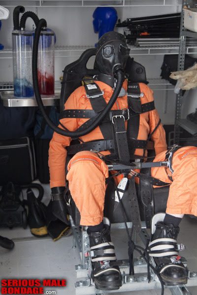 A person wearing an orange jumpsuit and gas mask is strapped securely into a mechanical restraint chair.