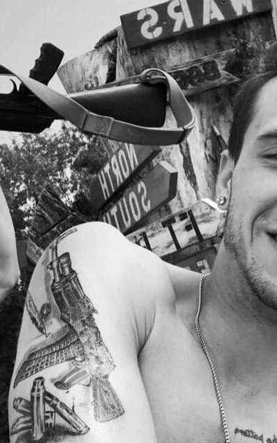 A shirtless, muscular man with a large firearm tattoo on his shoulder smiles outdoors near a rifle and directional signs.