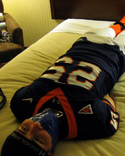 A man wearing a full football uniform is restrained on a bed with a tape gag and collar.