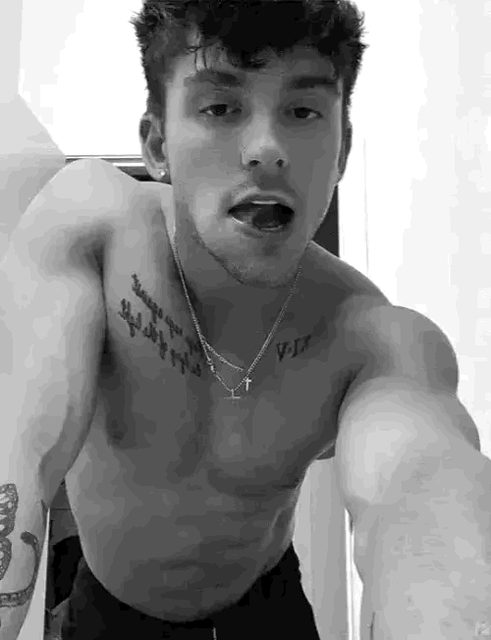 A young athletic man with chest tattoos leans toward the camera shirtless in a black and white portrait.