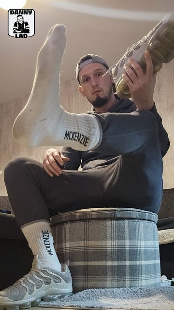 A man in a grey tracksuit and backwards cap poses with his feet in white McKenzie crew socks and Nike sneakers.