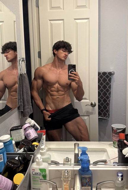 A muscular young man poses shirtless in gym shorts while taking a mirror selfie in a bathroom.