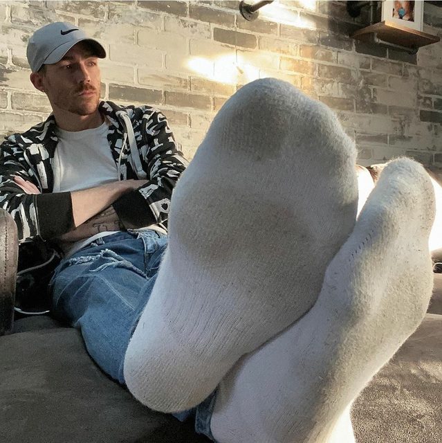 A man with a mustache and a baseball cap leans back on a sofa, presenting his white cotton socks to the camera.