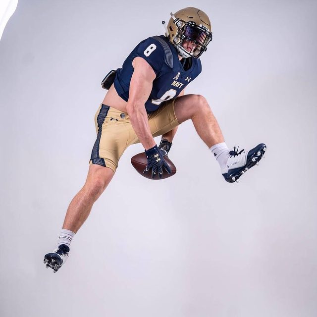 A muscular athlete in a full Navy football kit jumps into the air while holding a football in a studio setting.