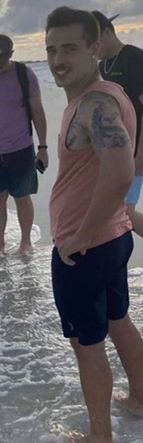 A tattooed young man wearing a pink tank top and blue swim shorts stands in the shallow water at a beach.