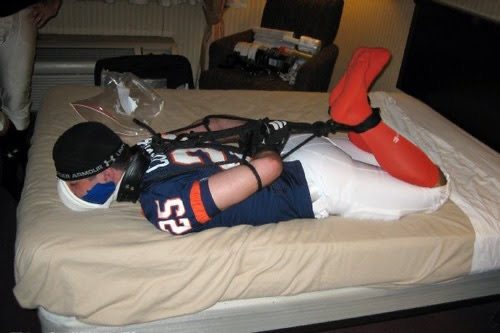 A bound male wearing a football jersey and orange socks lies hogtied face down on a bed.