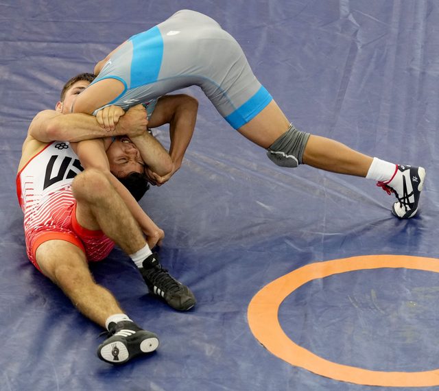 Two athletic young men in wrestling singlets are shown grappling and pinning each other on a blue competition mat.