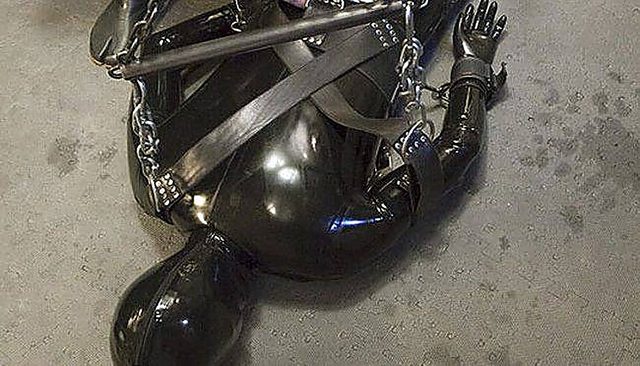 A person clad in a full black latex gimp suit and hood is bound on the floor with heavy straps, chains, cuffs, and a metal spreader bar.