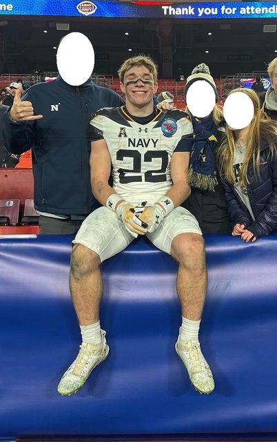 A muscular young football player in a dirty Navy jersey and white pants sits on a stadium barrier after a game.