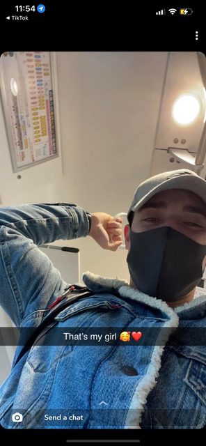 A young man in a denim jacket and baseball cap wearing a face mask holds a hand strap while riding public transport.