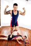A muscular tattooed man in a blue and red wrestling singlet flexes triumphantly while standing with one foot on his defeated opponent.