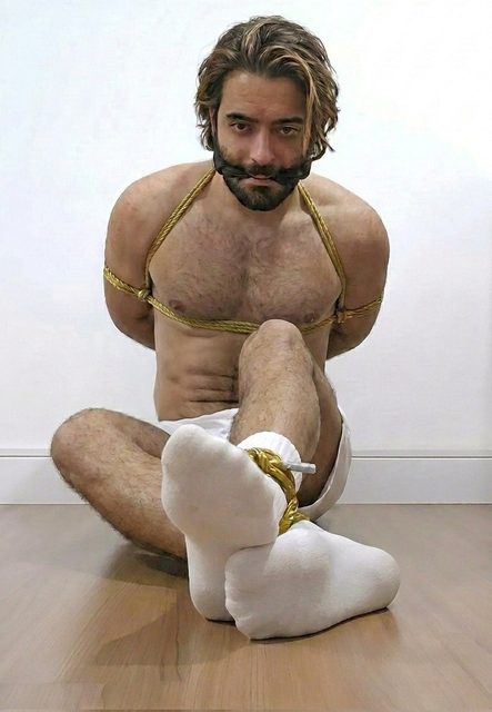 A bearded man sits on a wooden floor bound in gold rope with a gag in his mouth and his feet in white socks tied together.