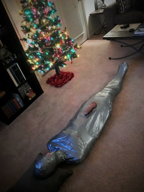 A person wrapped entirely in silver duct tape from head to toe lies restrained on the carpeted floor near a decorated Christmas tree with a dildo placed on their pelvis.