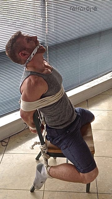 A muscular man wearing a grey tank top is bound tightly to a wooden chair with white rope and gagged with a bandana.
