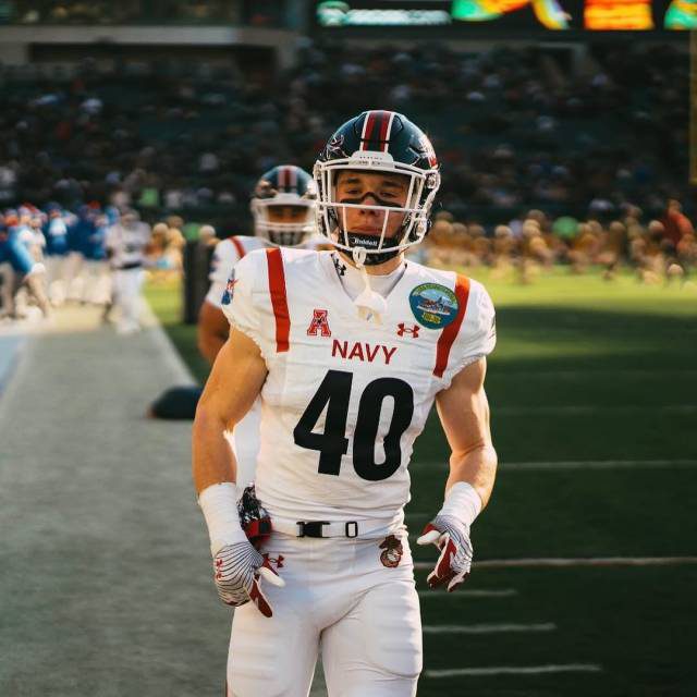 A muscular young football player in a full Navy uniform stands on the field wearing his helmet and gloves.