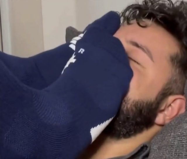 A bearded man lies back while a foot wearing a blue athletic sock is pressed firmly against his face.