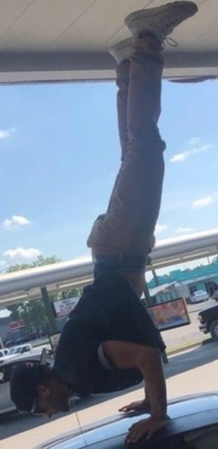 A fit man performs a handstand on top of a car roof in a public outdoor setting.