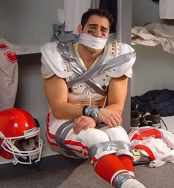A young football player wearing shoulder pads and athletic pants sits bound with duct tape and gagged in a locker room.
