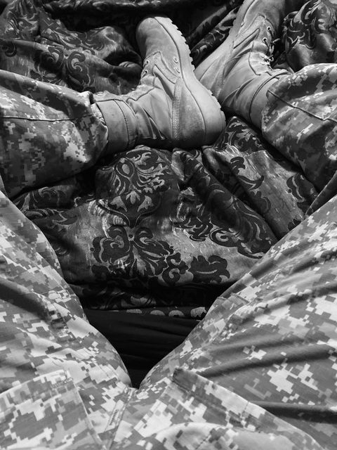 A point-of-view shot of a person in digital camouflage pants looking toward another set of legs in military fatigue pants and combat boots resting on a patterned blanket.