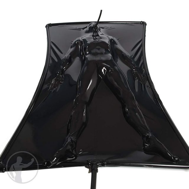 A muscular man is fully encased in a shiny black latex vacuum bed with a single breathing tube for air.