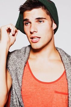 A young twunk wearing an orange tank top and a green beanie poses against a white background with a grey sweater over his shoulders.