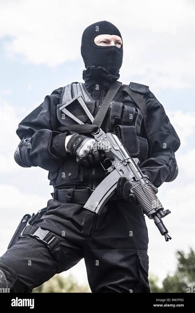 A man in full black tactical military gear, including a balaclava and vest, holds a rifle outdoors.