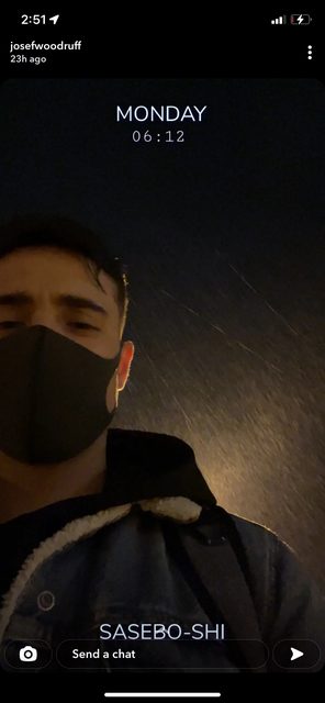 A young man wearing a face mask and a sherpa-lined denim jacket stands outdoors at night in the rain.