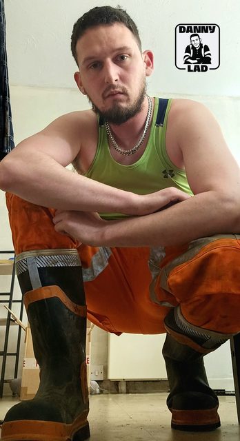 A bearded man in a lime green tank top and orange hi-vis work trousers squats while wearing heavy rubber boots.
