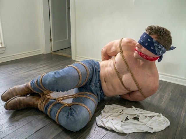 A muscular man in denim jeans and boots lies bound in natural rope with a bandana blindfold and gag.