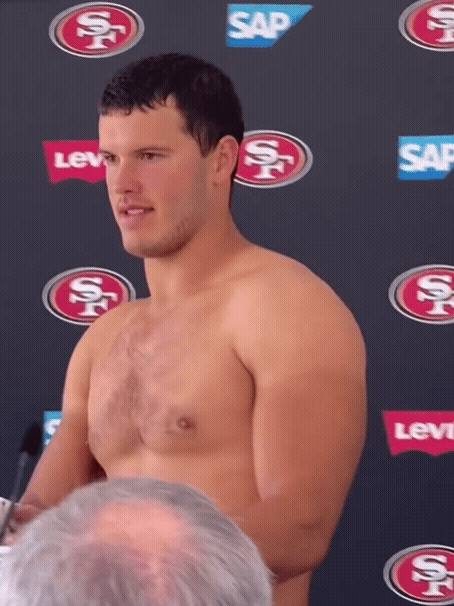 A shirtless, muscular man with light chest hair stands before a 49ers themed backdrop during a media event.
