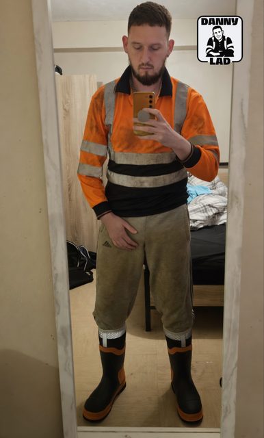 A bearded man takes a mirror selfie while wearing a dirty high-visibility work shirt, grey sweatpants, and rubber boots.