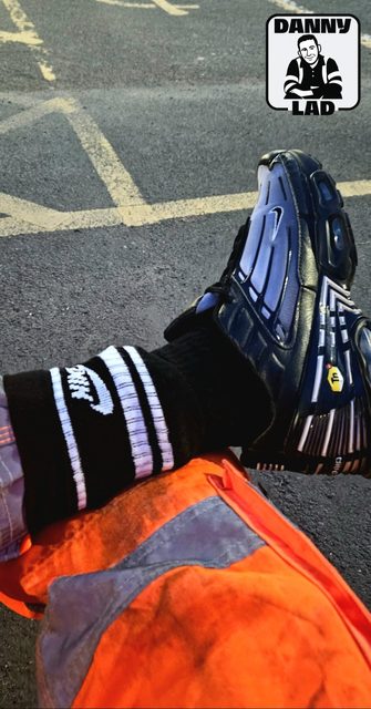 A seated person shows off their black Nike socks and blue TN sneakers while wearing hi-vis orange work trousers in an outdoor setting.
