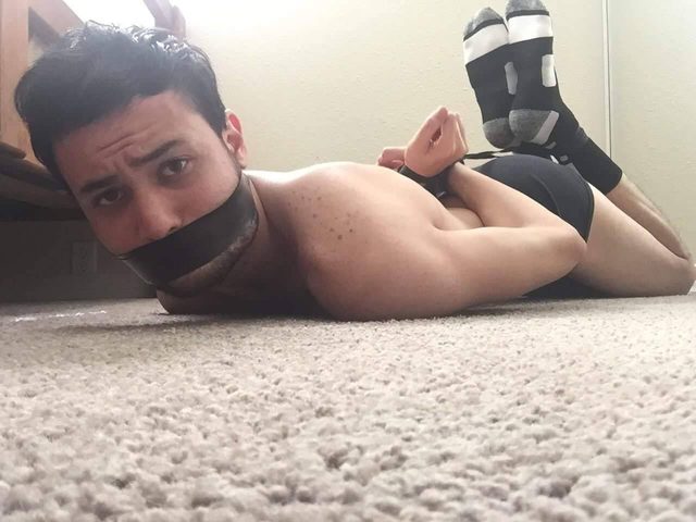 A submissive young man lies prone on a carpeted floor with his mouth taped and hands bound behind his back.