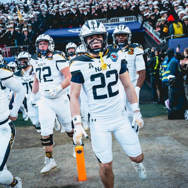 A group of muscular American football players in full padded uniforms and helmets walk onto the field at a stadium.