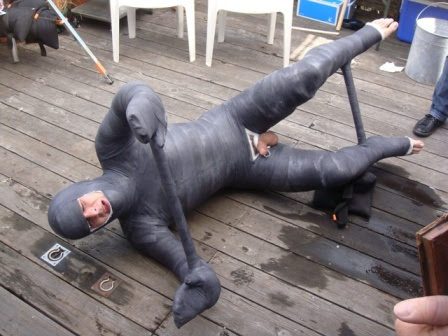 A man lies on a wooden deck, completely immobilized in a grey full-body cast with a cutout exposing his genitals.