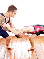 Two athletic men in wrestling singlets engage in a grappling match on a wooden floor.