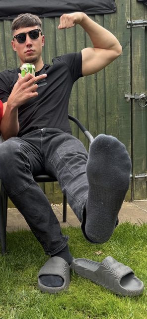 A young man sits outdoors, flexing his bicep and prominently displaying his feet in socks.