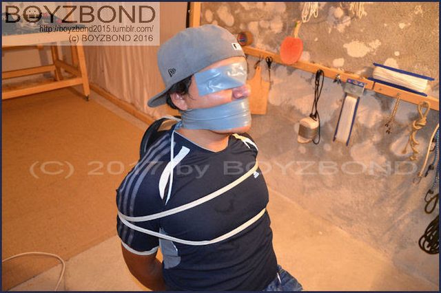 A young man wearing a grey baseball cap and athletic shirt is bound with rope, with duct tape covering his eyes and mouth in a basement setting.