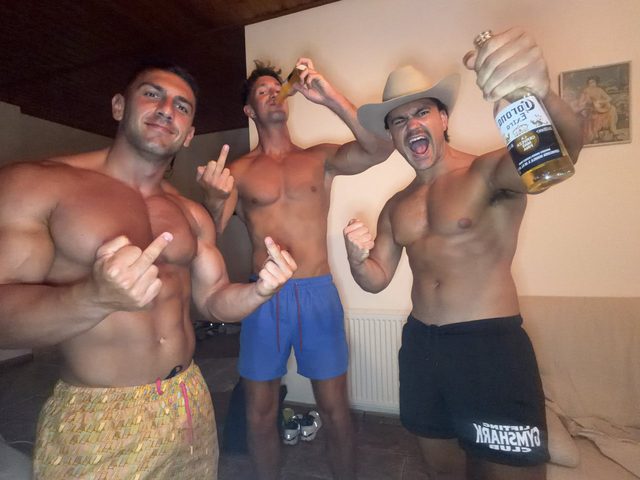 Three shirtless, muscular men in gym shorts and swim trunks drink beer and flex their physiques indoors.