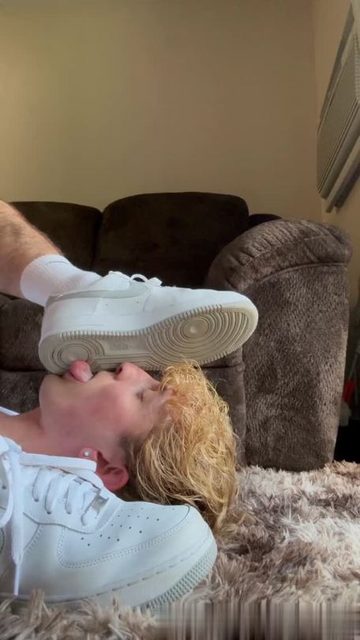 A person lies on a rug and licks the sole of a sneaker worn by another person.