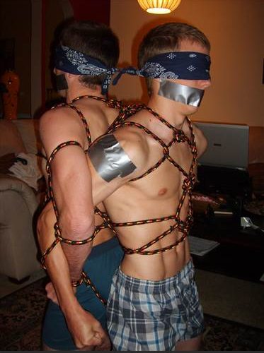 Two young men are bound back-to-back with patterned rope, blindfolded with bandanas, and gagged with duct tape indoors.