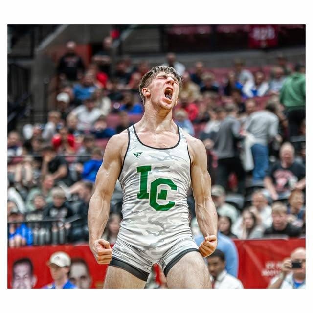 A muscular wrestler in a camouflage singlet flexes and yells in celebration inside a crowded arena.