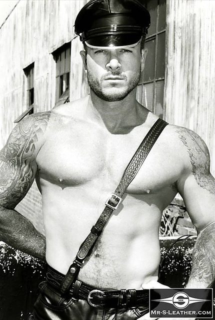 A shirtless muscular tattooed man wearing a leather muir cap and a leather crossbody strap poses outdoors.