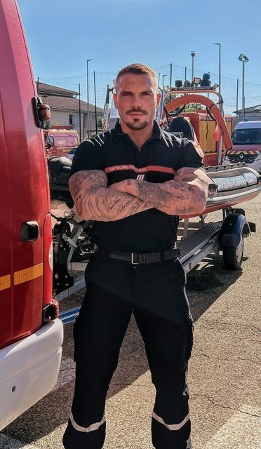 A muscular, tattooed man in a black Sapeurs-Pompiers firefighter uniform stands with crossed arms outdoors next to a fire truck and rescue boat.