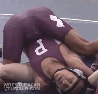 A wrestler in a maroon singlet and headgear pins an opponent on a mat, showcasing the tight fit of the spandex sportswear.