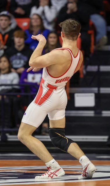 A fit young man in a tight white and red wrestling singlet stands on a mat in a gymnasium.