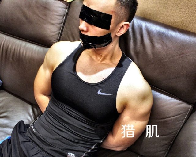 A muscular man wearing a black compression singlet is blindfolded and gagged with black tape while his arms are restrained behind his back on a leather sofa.