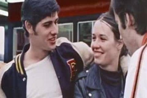 A young man wearing a classic varsity letterman jacket stands outdoors alongside friends in leather gear.