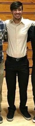 A smiling twink wearing a tan button-up shirt and black trousers stands in a gym.