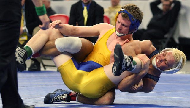 Two athletic male wrestlers wearing spandex singlets and protective headgear grapple intensely on a mat during a tournament match.